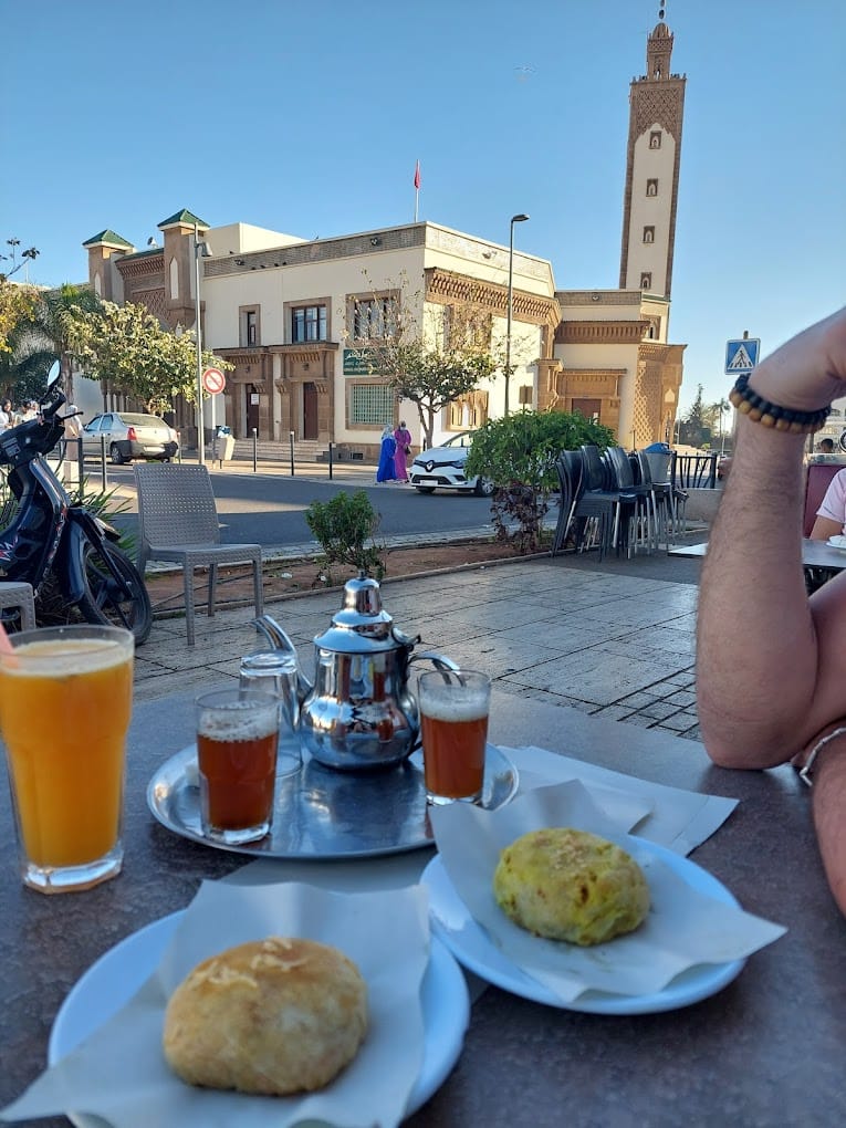 Talborjt Mosque in Agadir with a traditional Moroccan tea pot, tea glasses, and Moroccan sweets on a table in the foreground, showcasing Moroccan culture and hospitality